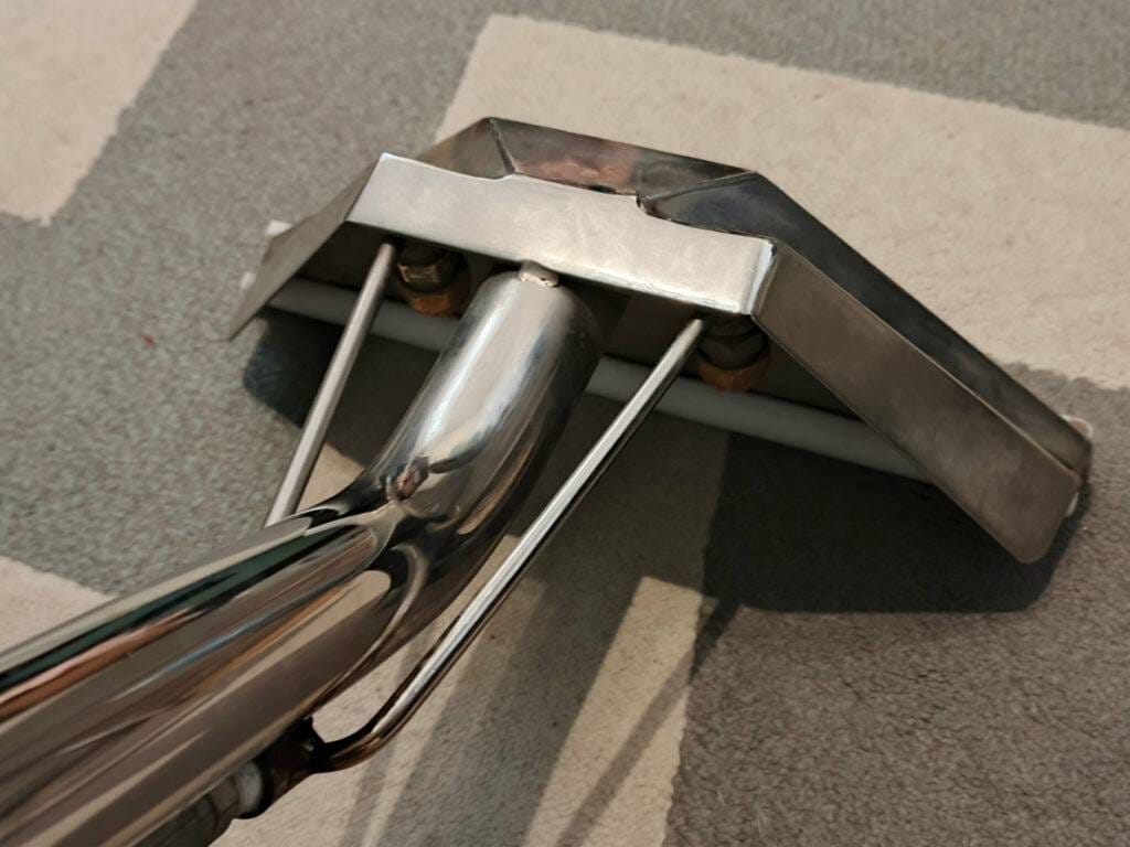 Zbliżenie na szeroką, stalową ssawkę ekstrakcyjną opartą o szary dywan - Podstawowe narzędzie, którego używa Turboszczotka, realizując mobilne pranie dywanów w Gdańsku.