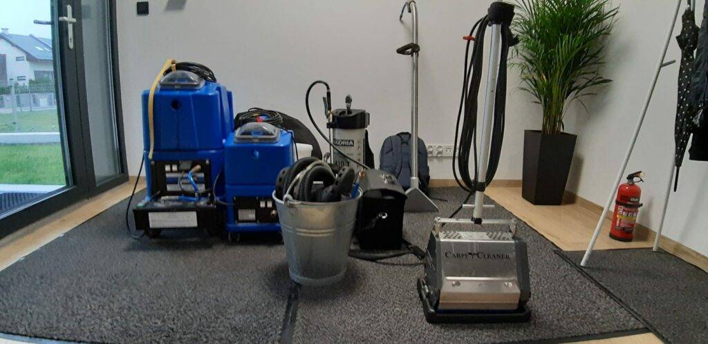 Profesjonalny sprzęt piorący, w tym odkurzacz ekstrakcyjny i szorowarka, rozstawione na szarym dywanie przed fachowym praniem dywanów w Gdyni.